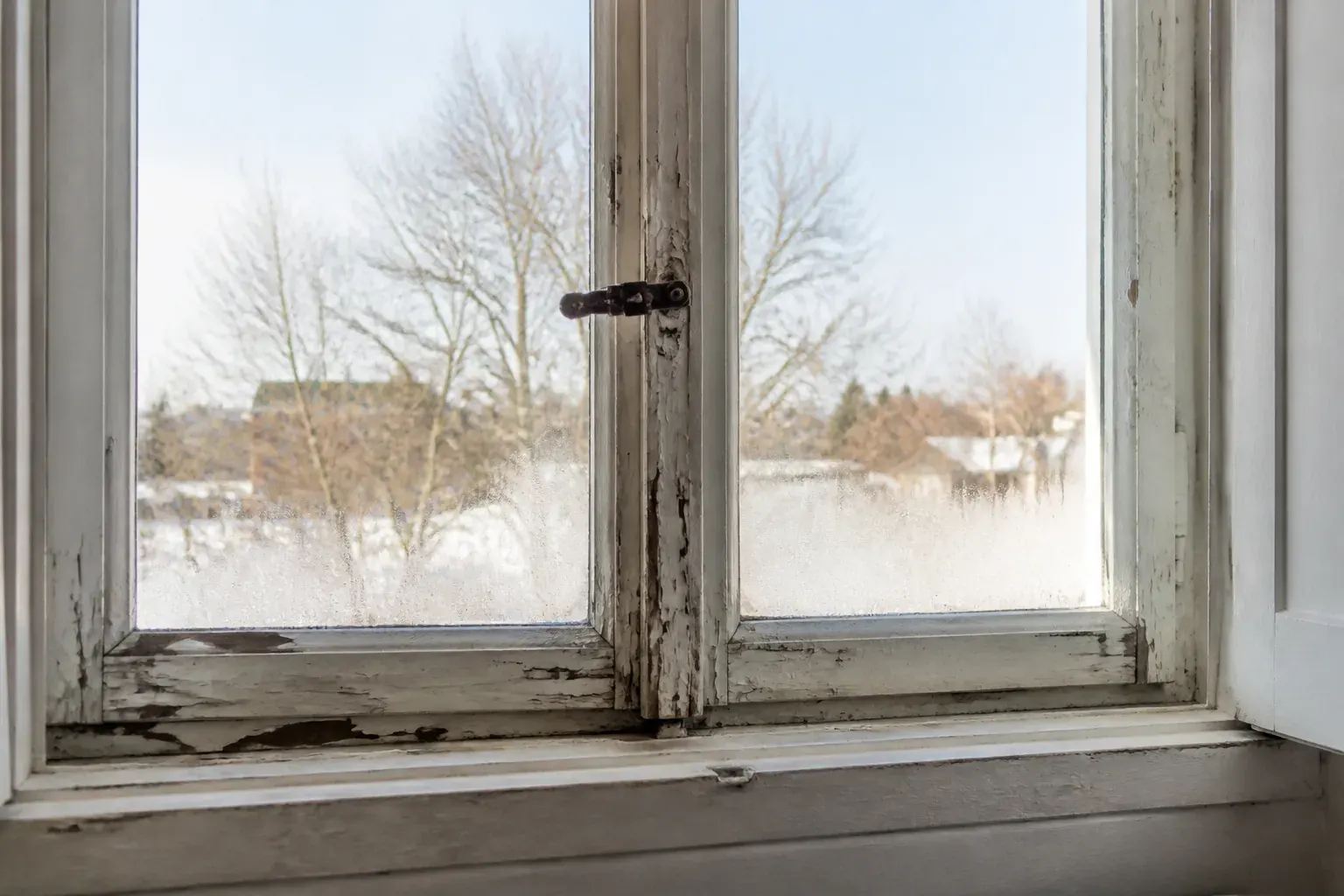 Ancienne fenêtre en bois à simple vitrage vue de l’intérieur, peinture écaillée sur le dormant et joint craquelé